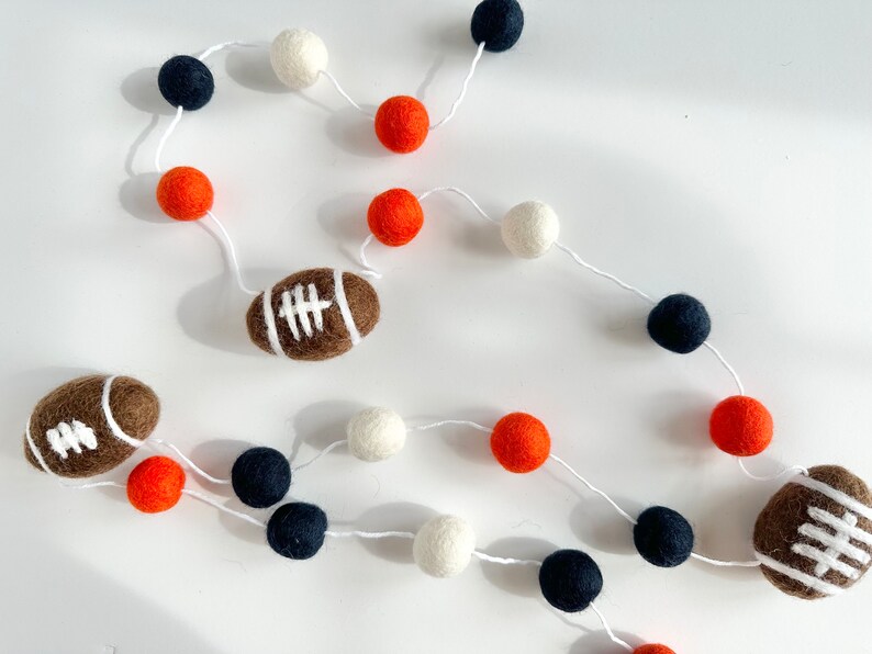 Puede incluir: Una guirnalda con bolas de fieltro blancas, naranjas y negras y dos balones de f&uacute;tbol de fieltro marrones.