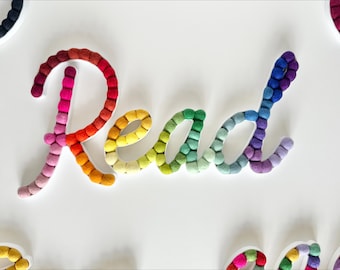 May include: The word "Read" spelled out in colorful felt pom-poms. The pom-poms are arranged in a rainbow pattern, starting with pink and red at the top and ending with purple and blue at the bottom.
