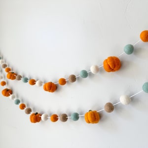 May include: A garland made of felt balls in orange, white, brown, and blue. The orange balls are shaped like pumpkins. The garland is strung on white string.