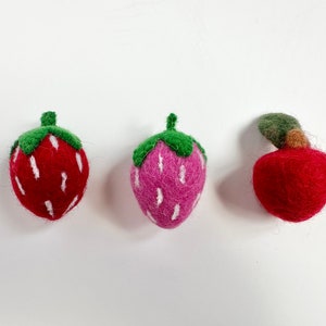 May include: Three felt fruit ornaments: a red strawberry, a pink strawberry, and a red apple. Each fruit has a green felt stem and leaves.