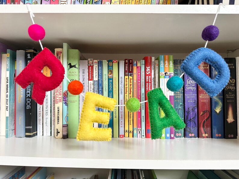 Puede incluir: Una pancarta de fieltro que deletrea "READ" en colores arco&iacute;ris con pompones colgando entre cada letra. La pancarta est&aacute; colgada frente a una estanter&iacute;a llena de libros.