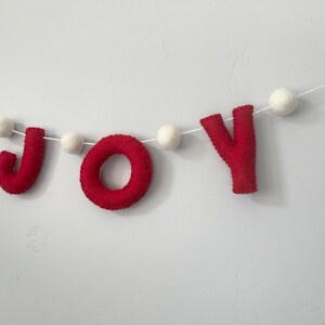 May include: A decorative garland featuring the word JOY spelled out in red felt letters, interspersed with small white felt balls. The letters and balls are strung together on a thin white cord, creating a horizontal banner for display.