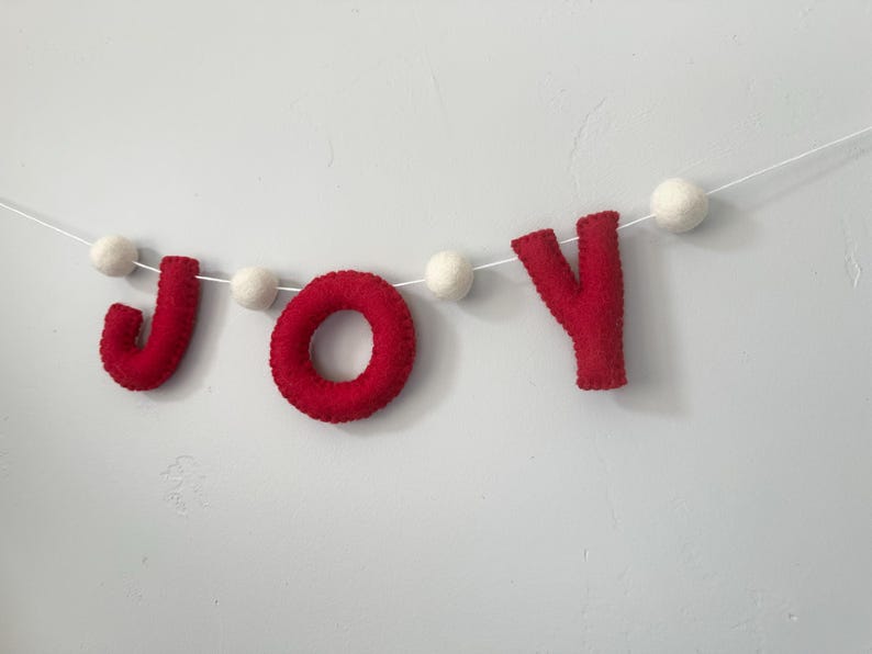 May include: A decorative garland featuring the word JOY spelled out in red felt letters, interspersed with small white felt balls. The letters and balls are strung together on a thin white cord, creating a horizontal banner for display.