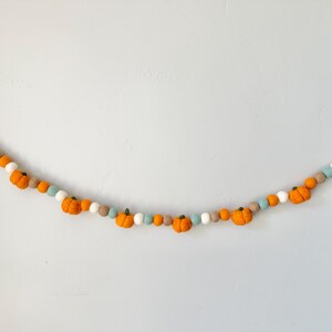 May include: A garland of felt pumpkins in orange, white, and light blue. The garland is strung together with a white string.