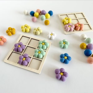 Pu&ograve; includere: Un tabellone di gioco in legno con un motivo a griglia e pezzi di fiori in feltro di vari colori, tra cui rosa, viola, blu, verde e giallo. I fiori sono disposti sul tabellone e sparsi intorno ad esso.
