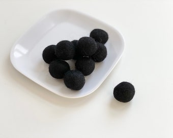 May include: A white ceramic plate with a pile of black felt balls. One black felt ball is sitting on a white surface next to the plate.