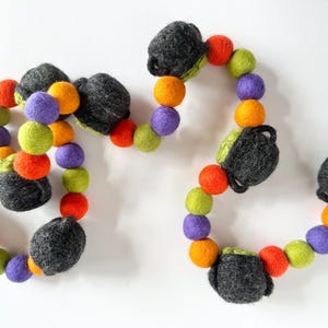 May include: A Halloween garland made of felt balls in orange, green, purple, and gray. The gray balls are shaped like cauldrons.