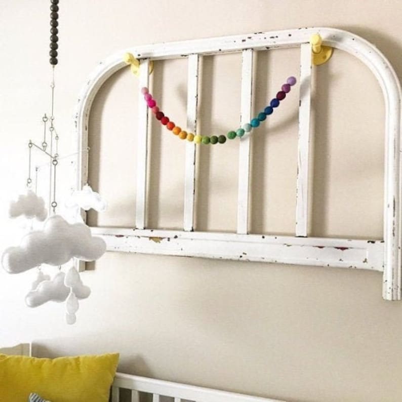 May include: A rainbow felt ball garland hangs from a white painted headboard with a decorative arch. The garland is made of colorful felt balls strung together on a white string. The headboard is attached to the wall with two yellow hooks.