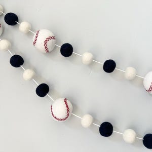 May include: A white and black felt ball garland with baseballs and white pom poms. The baseballs have red stitching.