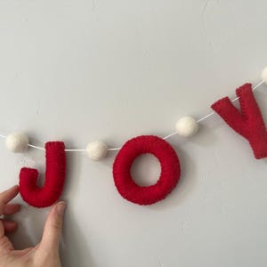 May include: A felt garland featuring the red letters J, O, and Y strung on a white cord, interspersed with small white felt balls. The letters are crafted from red felt material, with the letter O shaped as a circular ring and the J and Y in a block font style.