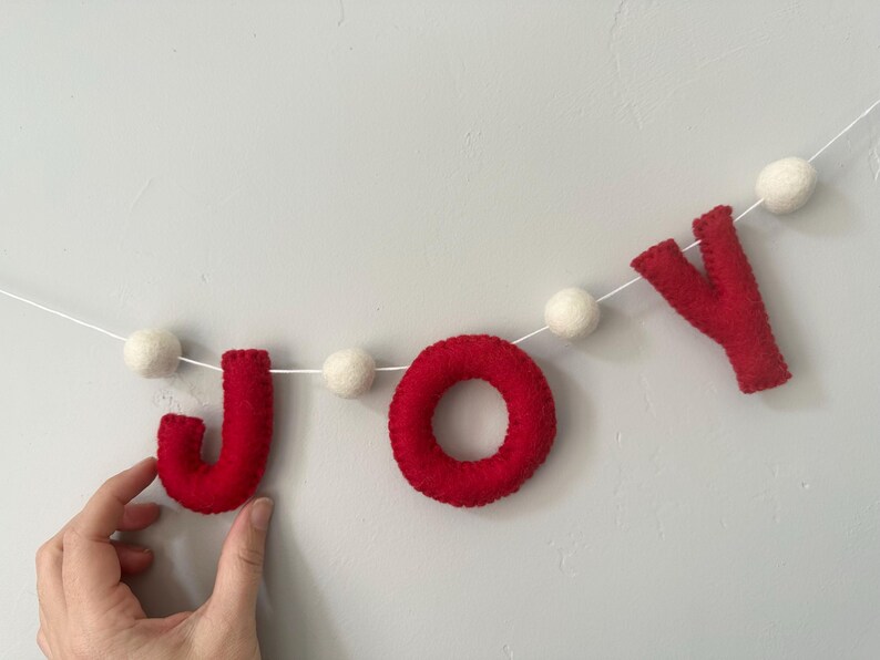 May include: A felt garland featuring the red letters J, O, and Y strung on a white cord, interspersed with small white felt balls. The letters are crafted from red felt material, with the letter O shaped as a circular ring and the J and Y in a block font style.