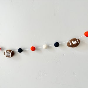 Puede incluir: Una guirnalda de fieltro con pompones de color naranja, blanco y azul marino alternados y balones de f&uacute;tbol marrones con costuras blancas.