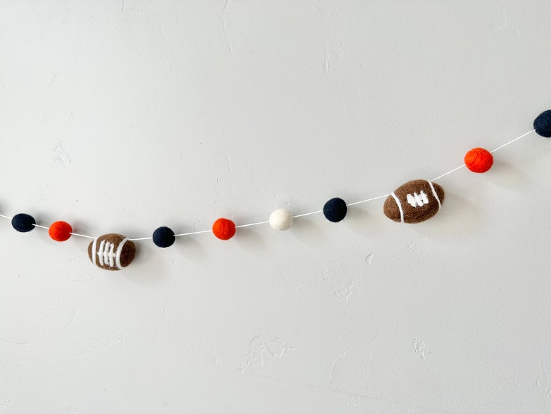 Puede incluir: Una guirnalda de fieltro con pompones de color naranja, blanco y azul marino alternados y balones de f&uacute;tbol marrones con costuras blancas.