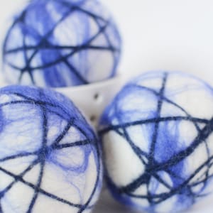 May include: Three white and blue felted wool balls with dark blue thread wrapped around them. The balls have a marbled blue pattern and are arranged in a white ceramic bowl. The balls are a decorative item.