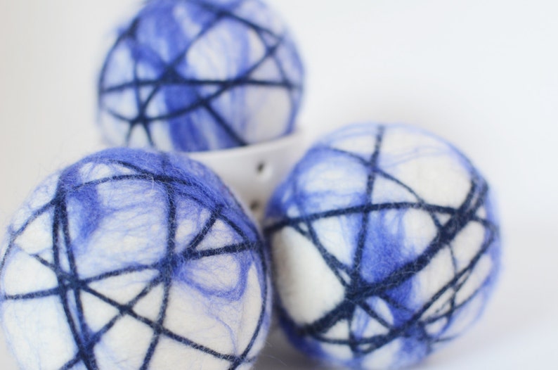 May include: Three white and blue felted wool balls with dark blue thread wrapped around them. The balls have a marbled blue pattern and are arranged in a white ceramic bowl. The balls are a decorative item.