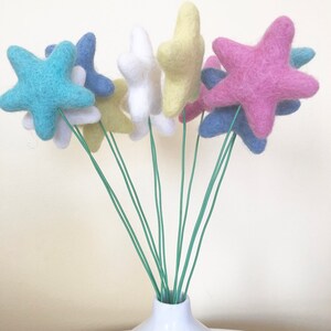 May include: A bouquet of felt star-shaped flowers in a white vase. The flowers are in shades of blue, pink, yellow, and white.