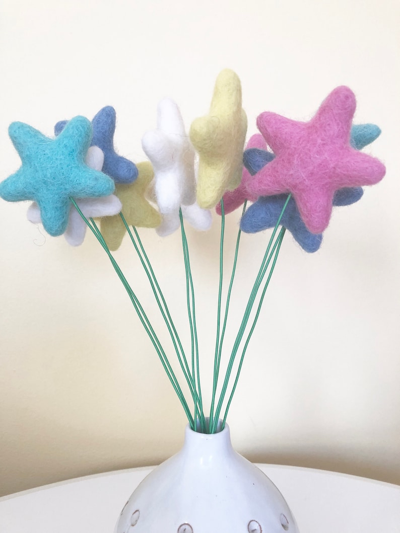 May include: A bouquet of felt star-shaped flowers in a white vase. The flowers are in shades of blue, pink, yellow, and white.