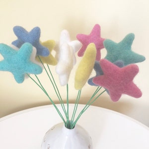 May include: A bouquet of felt star-shaped flowers in a white vase. The flowers are in shades of blue, green, yellow, pink, and white.