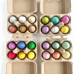 May include: Four open cardboard egg cartons containing a collection of twenty-four small, round, felted wool balls in a variety of solid colors including blue, white, cream, brown, green, pink, purple, yellow, red, and peach tones.