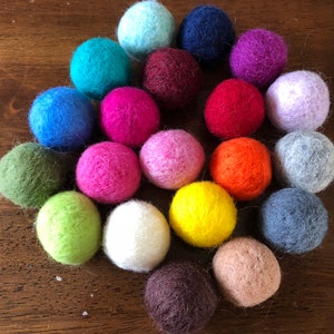 May include: A collection of 20 colorful felt balls in various shades of blue, green, red, purple, pink, orange, brown, and white. The balls are arranged in a circle on a wooden surface.