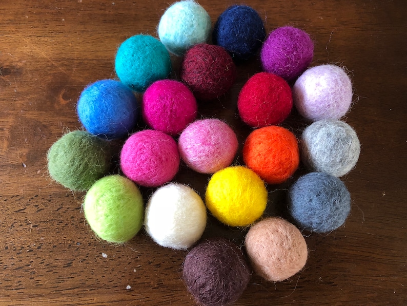 May include: A collection of 20 colorful felt balls in various shades of blue, green, red, purple, pink, orange, brown, and white. The balls are arranged in a circle on a wooden surface.