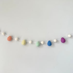 May include: A pastel rainbow felt garland with white pom poms. The garland features felt eggs in shades of purple, pink, green, yellow, blue, and light blue.