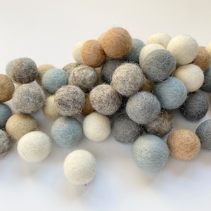 May include: A collection of felt balls in various shades of gray, blue, and beige. The balls are arranged in a pile, creating a soft and textured visual.