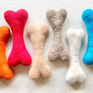 May include: Six felt dog chew toys in the shape of bones. The bones are in the colors orange, red, beige, gray, white, and blue.