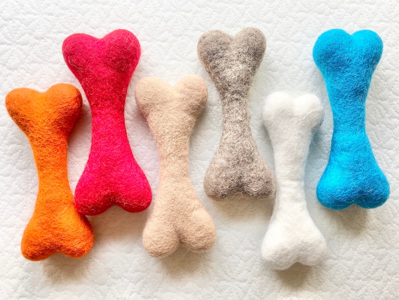 May include: Six felt dog chew toys in the shape of bones. The bones are in the colors orange, red, beige, gray, white, and blue.