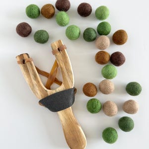 K&ouml;nnte beinhalten: Ein Holzschleuder mit schwarzem Griff und braunen Gummib&auml;ndern, zusammen mit einer Auswahl an bunten Filzkugeln in Gr&uuml;n-, Braun- und Beiget&ouml;nen. Die Filzkugeln haben einen Durchmesser von etwa 2,5 cm. Auf wei&szlig;em Hintergrund angeordnet.