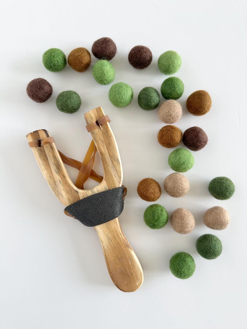 K&ouml;nnte beinhalten: Ein Holzschleuder mit schwarzem Griff und braunen Gummib&auml;ndern, zusammen mit einer Auswahl an bunten Filzkugeln in Gr&uuml;n-, Braun- und Beiget&ouml;nen. Die Filzkugeln haben einen Durchmesser von etwa 2,5 cm. Auf wei&szlig;em Hintergrund angeordnet.