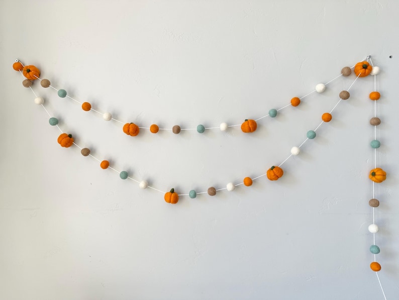 May include: A garland with felt orange pumpkins and white, gray, and brown pom poms. The garland is strung on white thread.