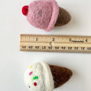 Pode incluir: Dois cones de gelado de feltro. Um &eacute; rosa com uma cereja vermelha no topo. O outro &eacute; branco com granulado colorido e um cone castanho. Uma r&eacute;gua est&aacute; no fundo.