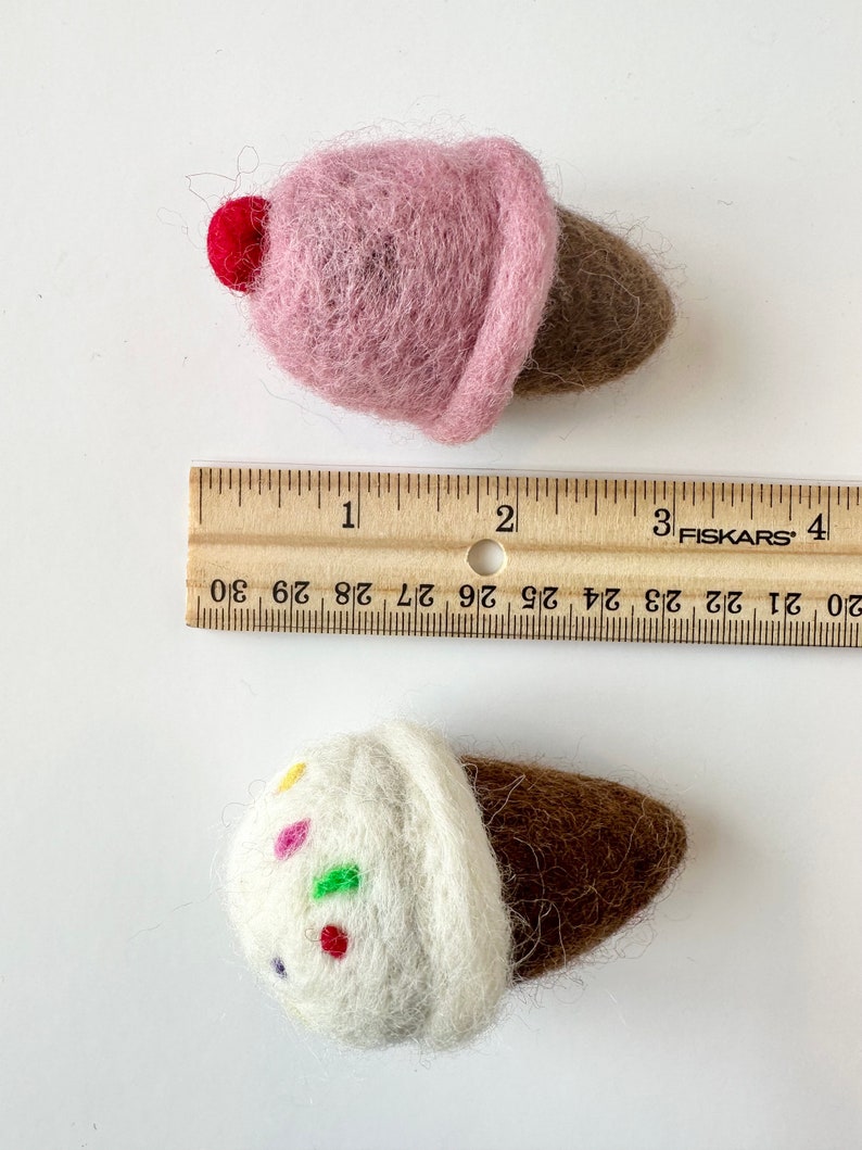 Pode incluir: Dois cones de gelado de feltro. Um &eacute; rosa com uma cereja vermelha no topo. O outro &eacute; branco com granulado colorido e um cone castanho. Uma r&eacute;gua est&aacute; no fundo.