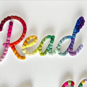 May include: The word Read is spelled out in cursive script using small, round, felted wool balls. The letters feature a vibrant gradient color scheme, transitioning from shades of red and pink to yellow, green, blue, and purple across the word.