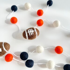 Puede incluir: Una guirnalda de cuerda blanca con bolas de fieltro naranja, blancas y azul marino. Dos balones de f&uacute;tbol de fieltro marr&oacute;n est&aacute;n incluidos en la guirnalda.