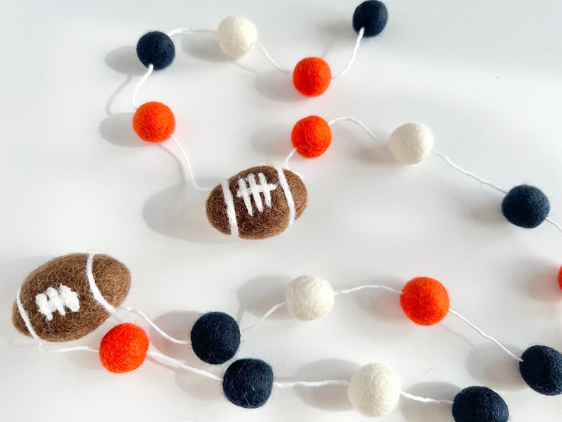 Puede incluir: Una guirnalda de cuerda blanca con bolas de fieltro naranja, blancas y azul marino. Dos balones de f&uacute;tbol de fieltro marr&oacute;n est&aacute;n incluidos en la guirnalda.
