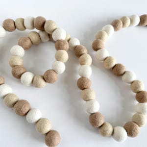 May include: A garland of felt balls in beige, cream, and white colors. The garland is made of individual felt balls strung together.