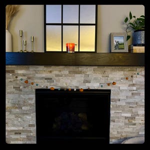 May include: A fireplace mantel with a stone surround and a garland of small pumpkins and white pom poms. There are three gold candle holders on the mantel and a lit candle in a glass jar.