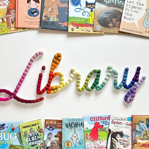 May include: A wall-mounted sign spelling the word Library in cursive script, constructed from a series of small, multi-colored felt balls in a gradient pattern ranging from pink and red to yellow, green, blue, and purple, positioned above and below children's books.