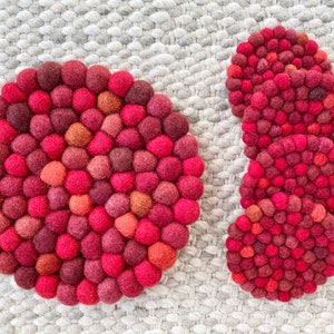 May include: Four red and burgundy felt coasters made of small, round balls. The coasters are arranged in a stack with the top coaster slightly offset from the bottom coasters.