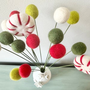 May include: A white vase with a floral design holds a bouquet of felt balls in red, green, and white. The felt balls are arranged in a whimsical, festive style, resembling candy canes and ornaments.