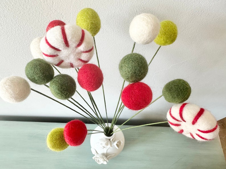 May include: A white vase with a floral design holds a bouquet of felt balls in red, green, and white. The felt balls are arranged in a whimsical, festive style, resembling candy canes and ornaments.