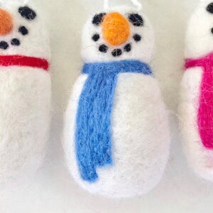 May include: Three felt snowman ornaments with different colored scarves. The snowman on the left has a red scarf, the middle snowman has a blue scarf, and the snowman on the right has a pink scarf.