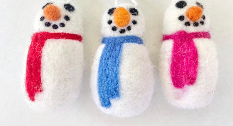 May include: Three felt snowman ornaments with different colored scarves. The snowman on the left has a red scarf, the middle snowman has a blue scarf, and the snowman on the right has a pink scarf.