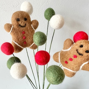 May include: A festive arrangement of felt gingerbread people and colorful pom-poms. The gingerbread people are brown with white icing and red buttons. The pom-poms are red, white, and green.