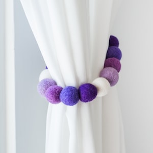 May include: A white curtain with a purple and white pom-pom curtain tieback.