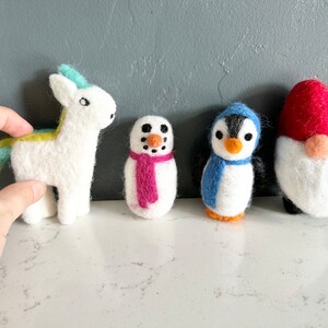 May include: Five felt animal ornaments: a white unicorn with a blue and yellow mane, a snowman with a pink scarf, a penguin with a blue scarf, and a red and white gnome.