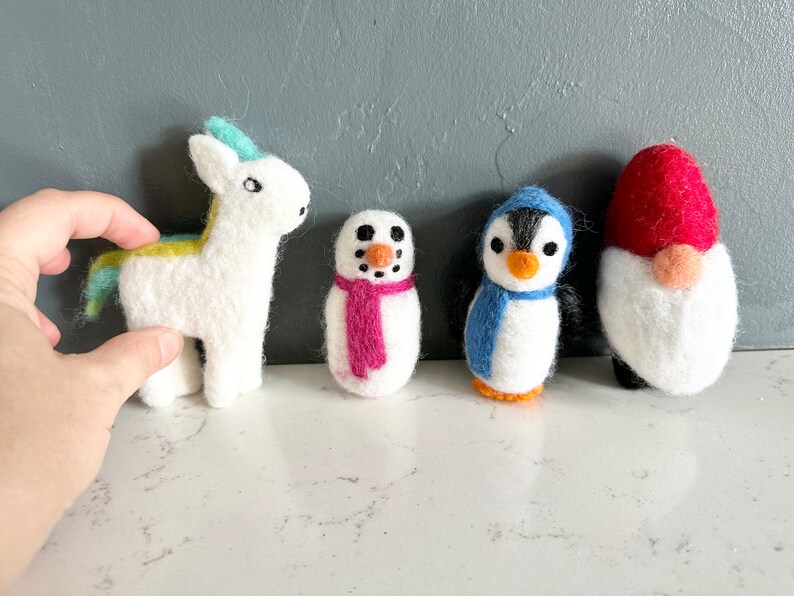May include: Five felt animal ornaments: a white unicorn with a blue and yellow mane, a snowman with a pink scarf, a penguin with a blue scarf, and a red and white gnome.