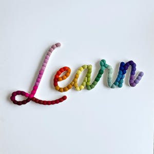 May include: The word Learn written in cursive script, constructed from small felt balls in a gradient of colors including dark red, pink, orange, yellow, green, teal, blue, and purple, arranged on a plain white background.